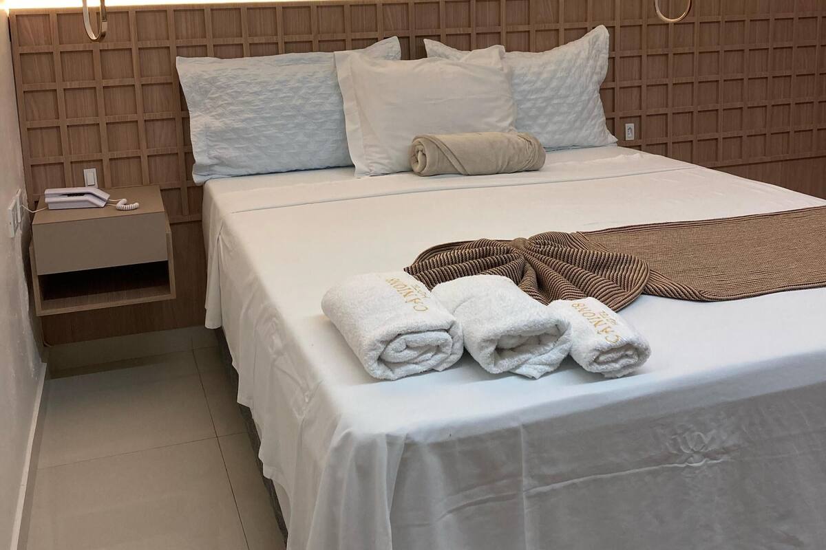 Un lit bien fait, avec du linge de lit blanc et deux serviettes enroulées, brodées du mot « Cannes », une tête de lit en bois, une table de chevet avec un téléphone et un livre, et un luminaire fixé au mur.