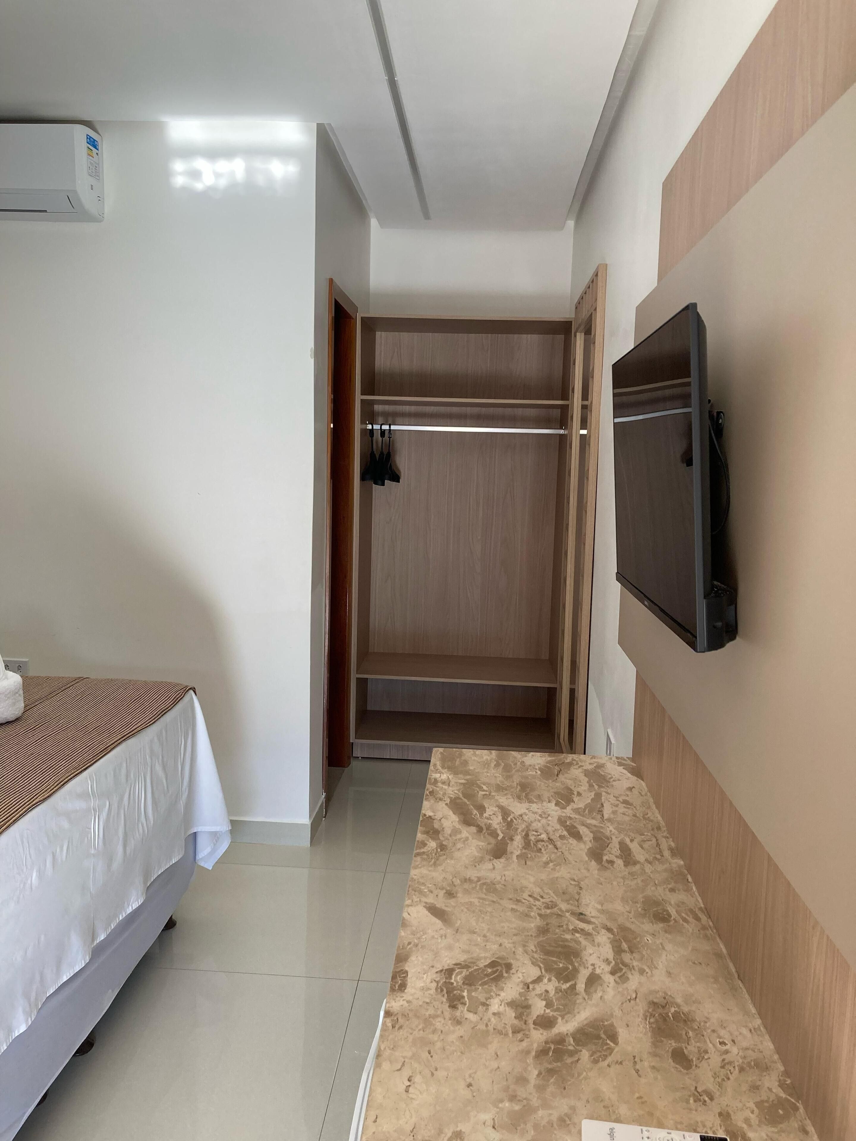 Una habitación de hotel con cama, televisión montada en la pared, armario de madera y suelo de baldosas.