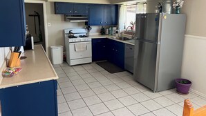 Cocina pequeña con estufa, lavamanos y refrigerador. Hay una ventana con cortinas, una planta en maceta y un basurero pequeño.