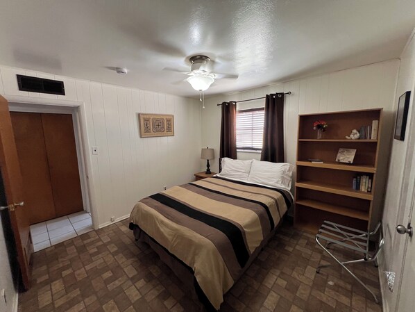 Un dormitorio con cama, mesita de noche, estantería, puerta y ventilador de techo.