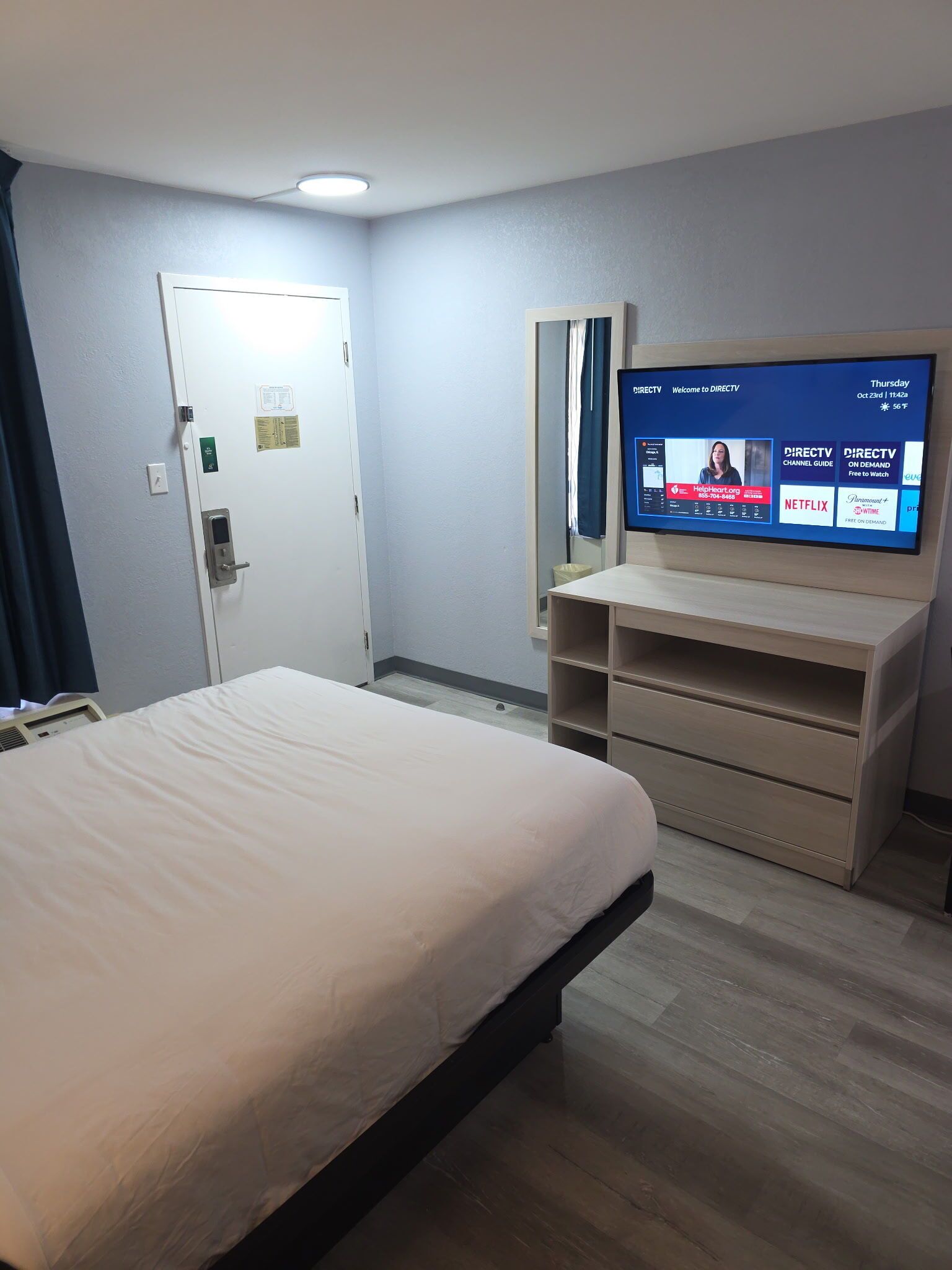 Habitación de hotel con cama, escritorio con monitor de computadora mostrando canales DIRECTV, y puerta con luz.