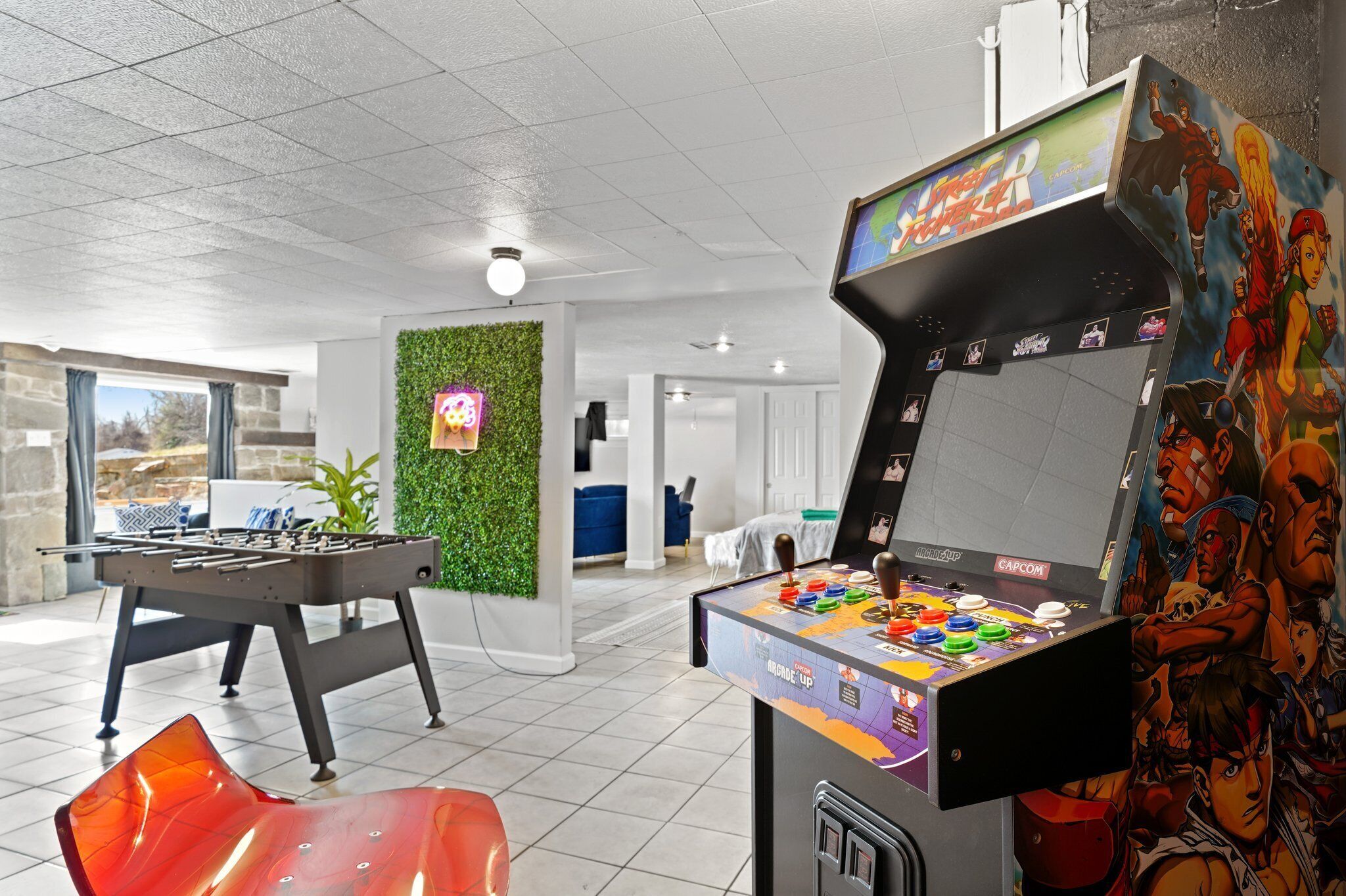 A room with a foosball table, a Capcom arcade machine, and a green wall with a neon sign.