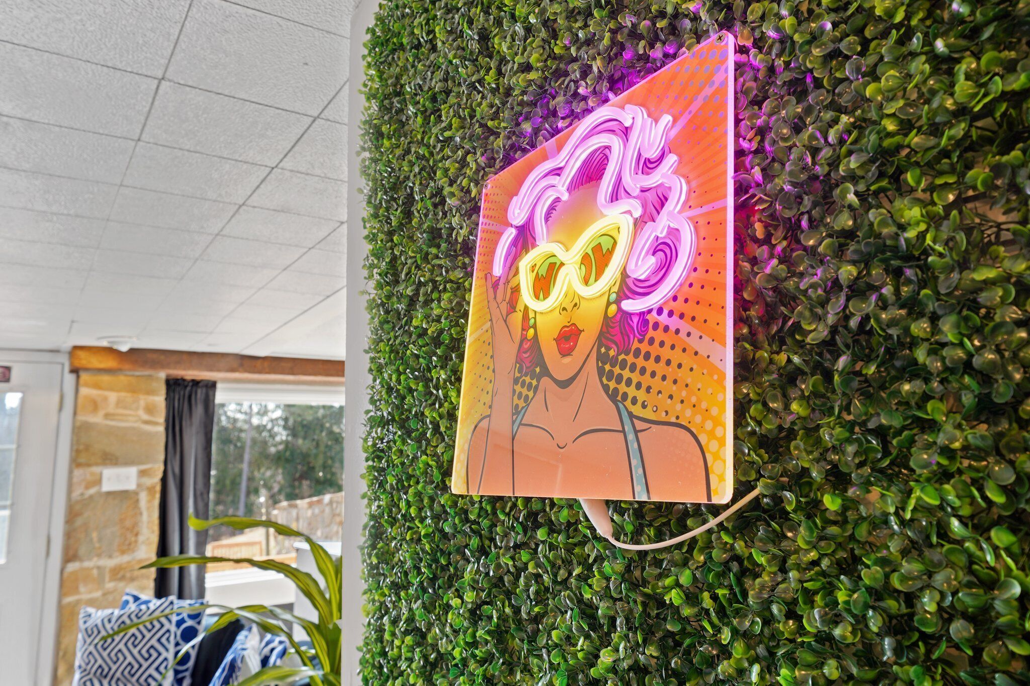Une enseigne au néon représentant une femme portant des lunettes de soleil et une robe, fixée à un mur sur lequel figure un décor de feuilles vertes.