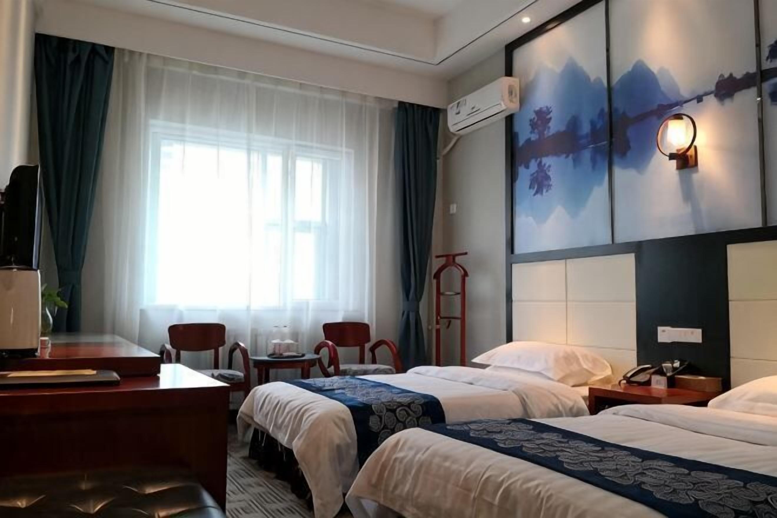 A hotel room with two beds, a desk with a chair, a small table, and a window with curtains.