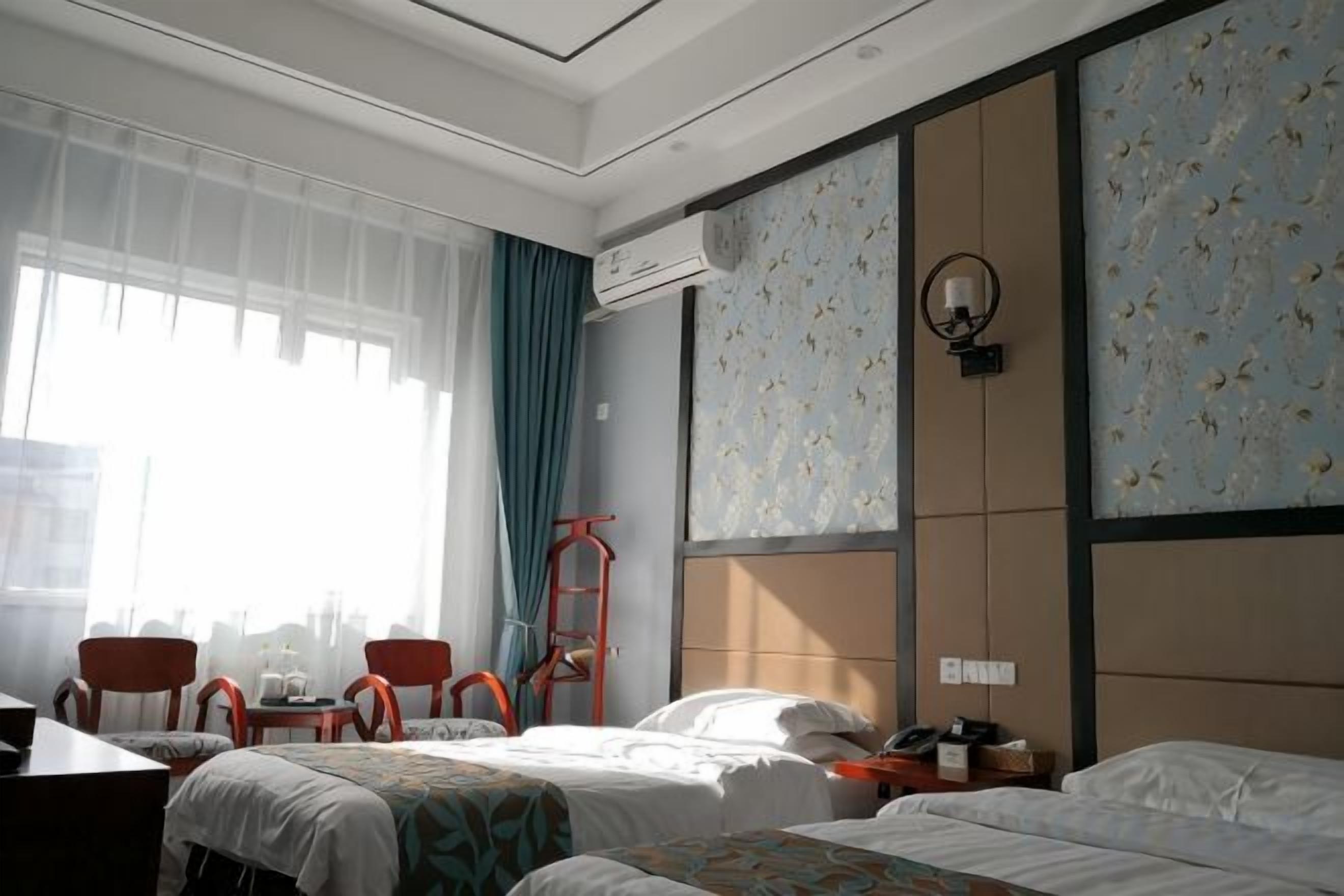 Een hotelkamer met een bed, een bureau en een stoel. Er is een raam met gordijnen, een wandairconditioner en een wandklok.