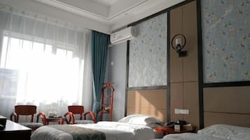 Een hotelkamer met een bed, een bureau en een stoel. Er is een raam met gordijnen, een wandairconditioner en een wandklok.