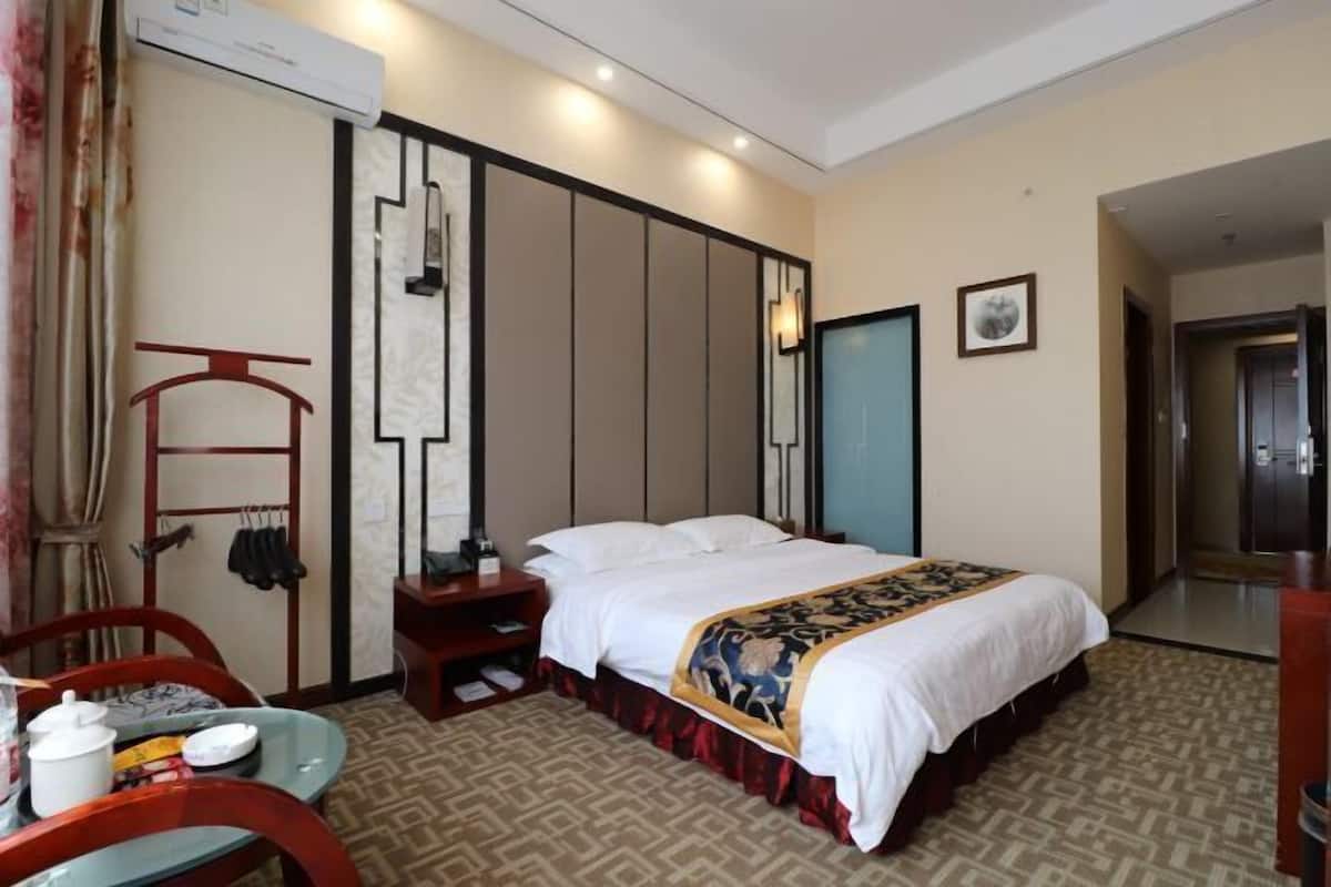 Habitación de hotel con una cama, una mesita con hervidor, una silla, perchero y una puerta que da a otra habitación.
