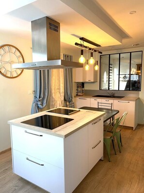 Une cuisine moderne dotée d’un îlot central, d’une hotte en acier inoxydable, de luminaires suspendus et d’un grand miroir au-dessus du plan de travail.