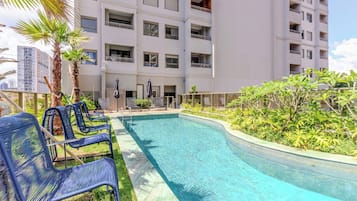 A swimming pool with lounge chairs, surrounded by greenery and a multi-story building with balconies.