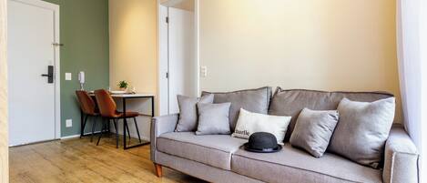 A living room with a grey sofa, a dining table with chairs, and a door leading to another room.