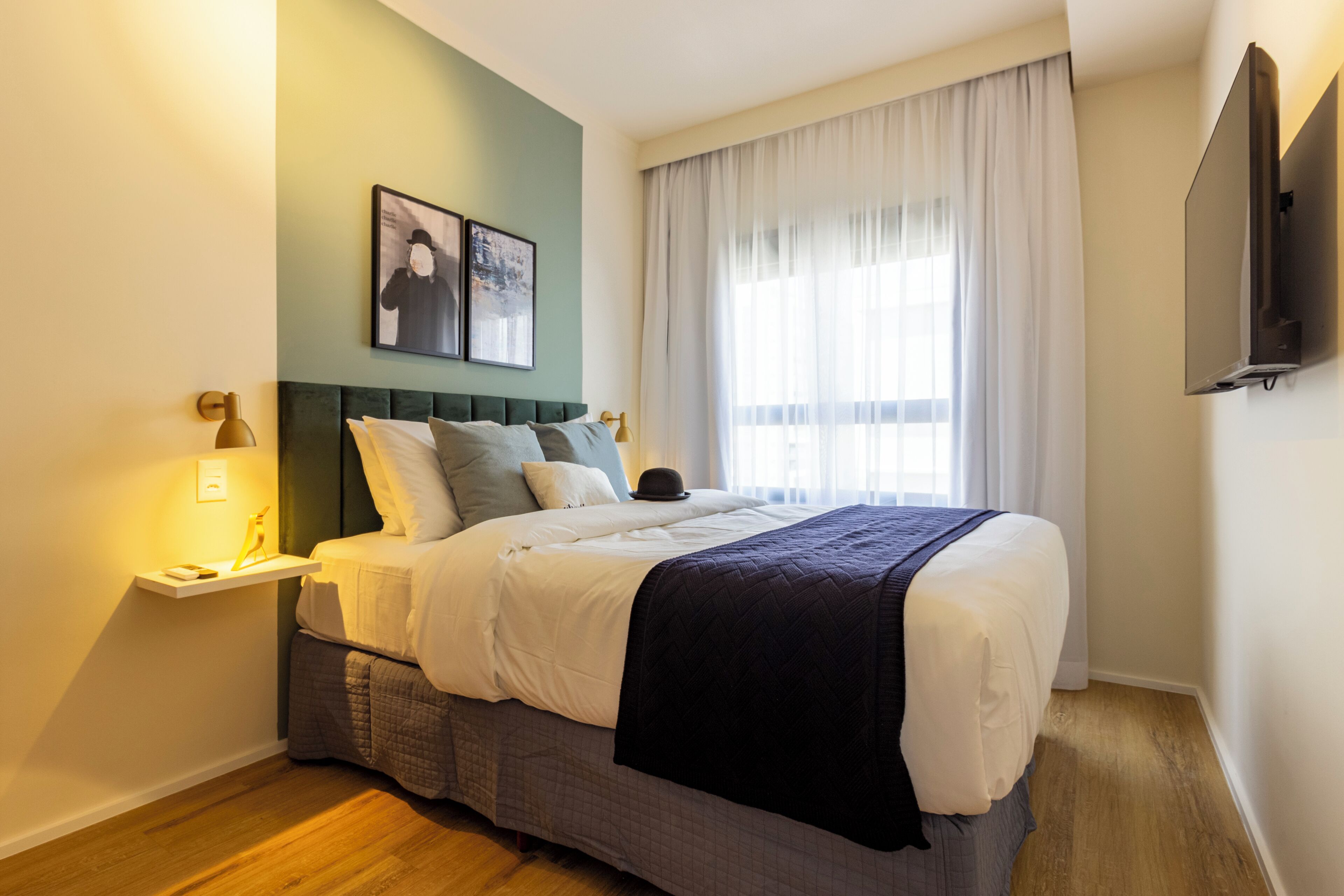 A hotel room with a bed, a bedside table, a wall-mounted TV, and a window with curtains.