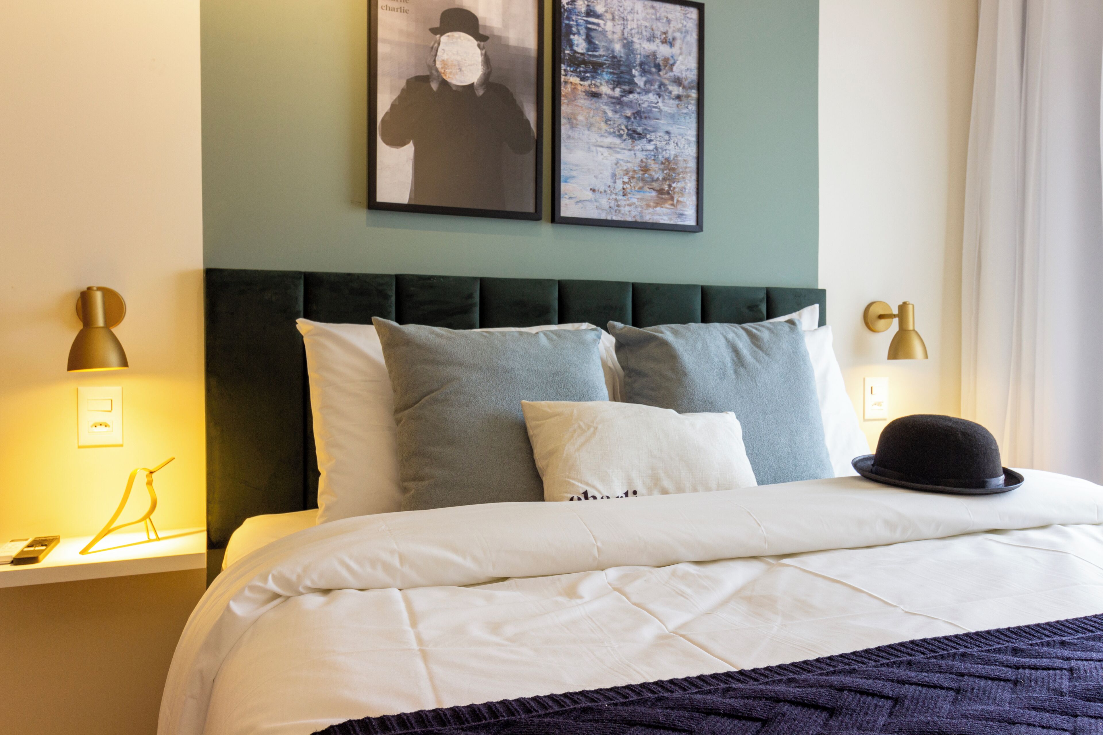 A neatly made bed with a dark headboard, white bedding, and a black hat on the pillow. There are two framed pictures on the wall above the bed.