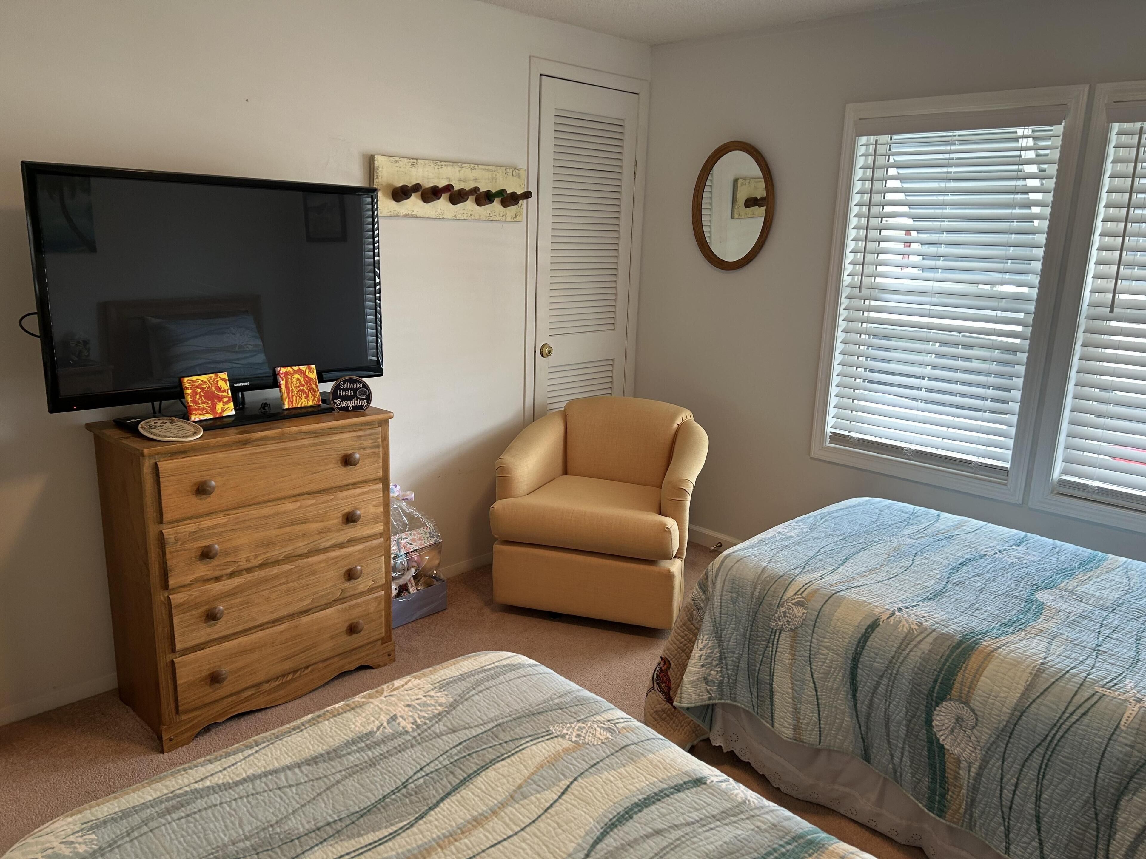 Ein Schlafzimmer mit einem Fernseher auf einer Holzkommode, einem beigen Sessel, zwei Betten mit gestreifter Bettwäsche, einem Fenster mit Rollläden und einer Tür mit Spiegel.