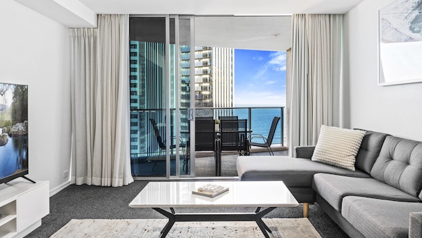 Una sala de estar moderna con un sofá gris, una mesa de centro blanca y un televisor de pantalla plana. Hay un balcón con vista al mar y un edificio al fondo.
