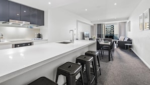 Una cocina moderna con encimera blanca, armarios oscuros y lavavajillas integrado. Hay tres taburetes en la encimera y una zona de comedor con mesa y sillas.
