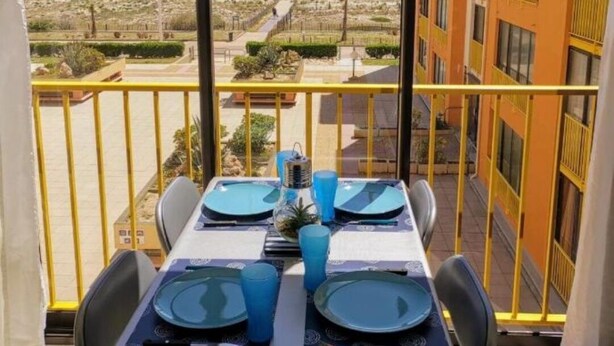 Mesa de comedor con platos y vasos azules, con vistas a la costa y edificios al fondo.