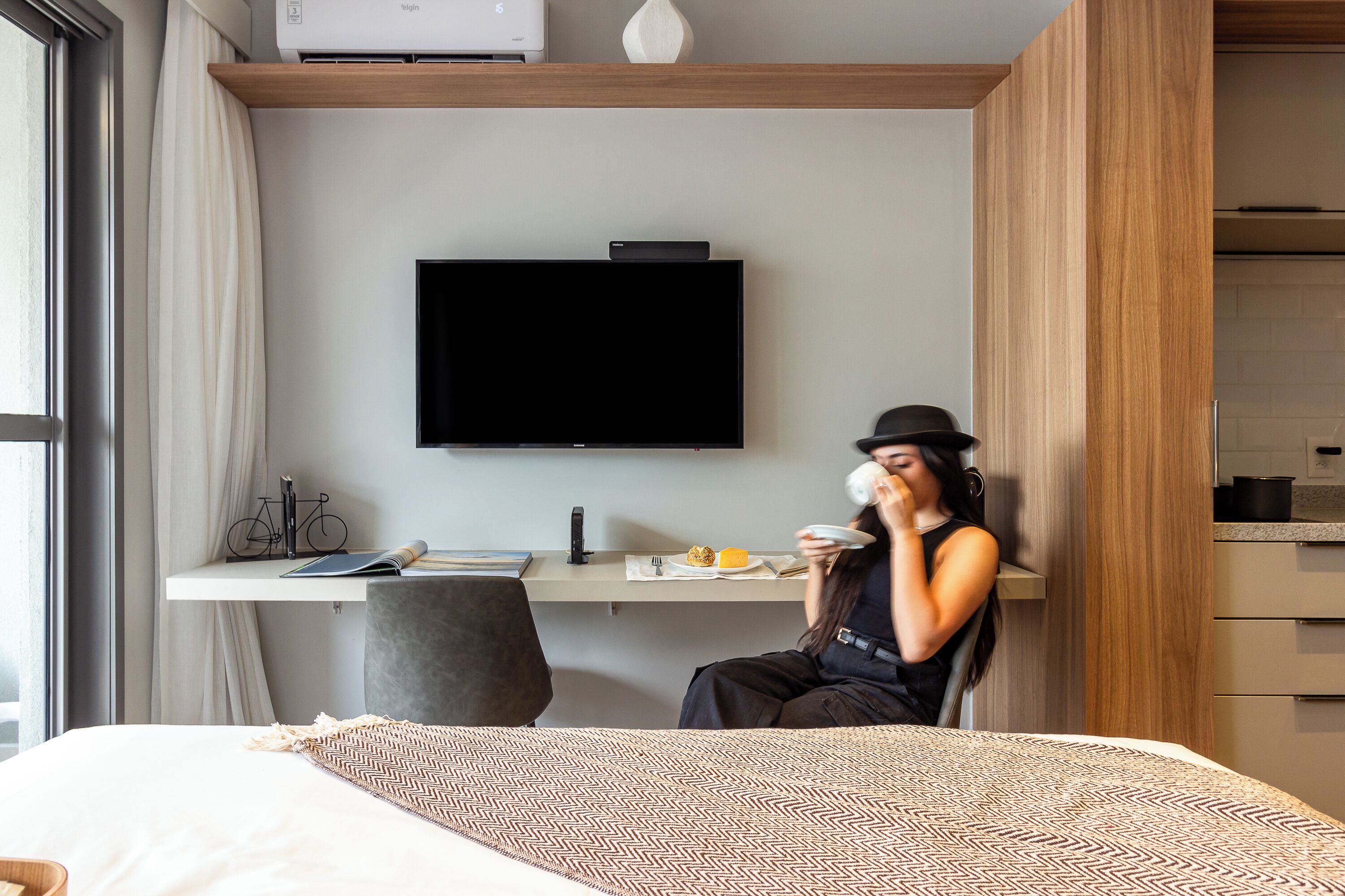 A modern hotel room with a bed, a desk with a flat-screen TV, a chair, and a dining area with a table and chairs.