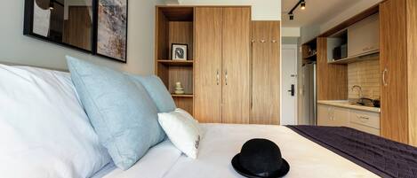 Une chambre à coucher avec un lit, des oreillers, un chapeau, une armoire et un coin cuisine.