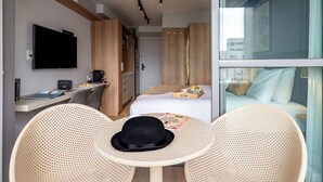 A modern hotel room with a flat-screen TV, a dining area with a table and chairs, a bed with a nightstand, and a window with a city view. - Charlie  Lumiere Klabin (São Paulo)