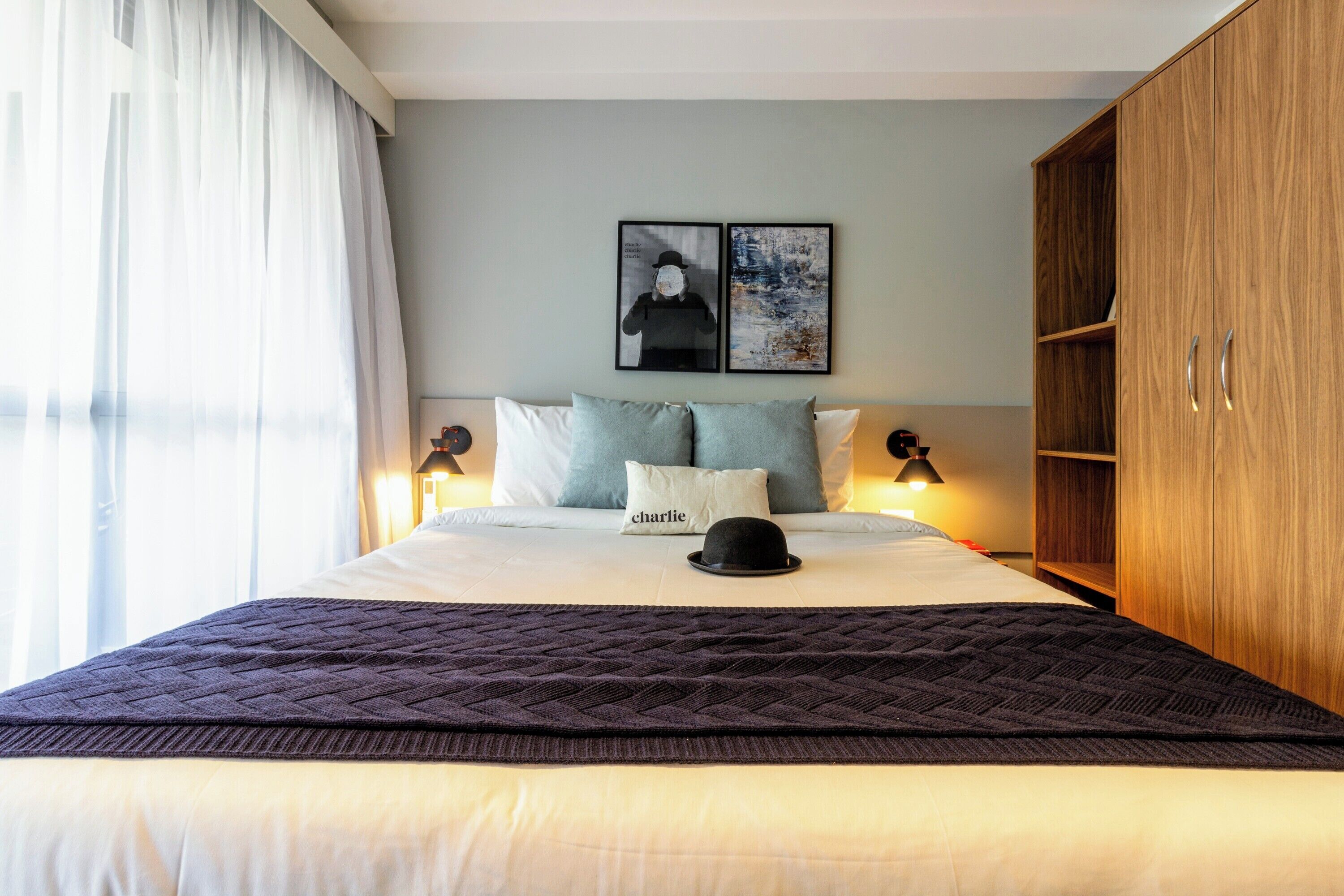 A neatly made bed with a quilted blanket, two pillows, a black hat, and a framed picture on the wall.