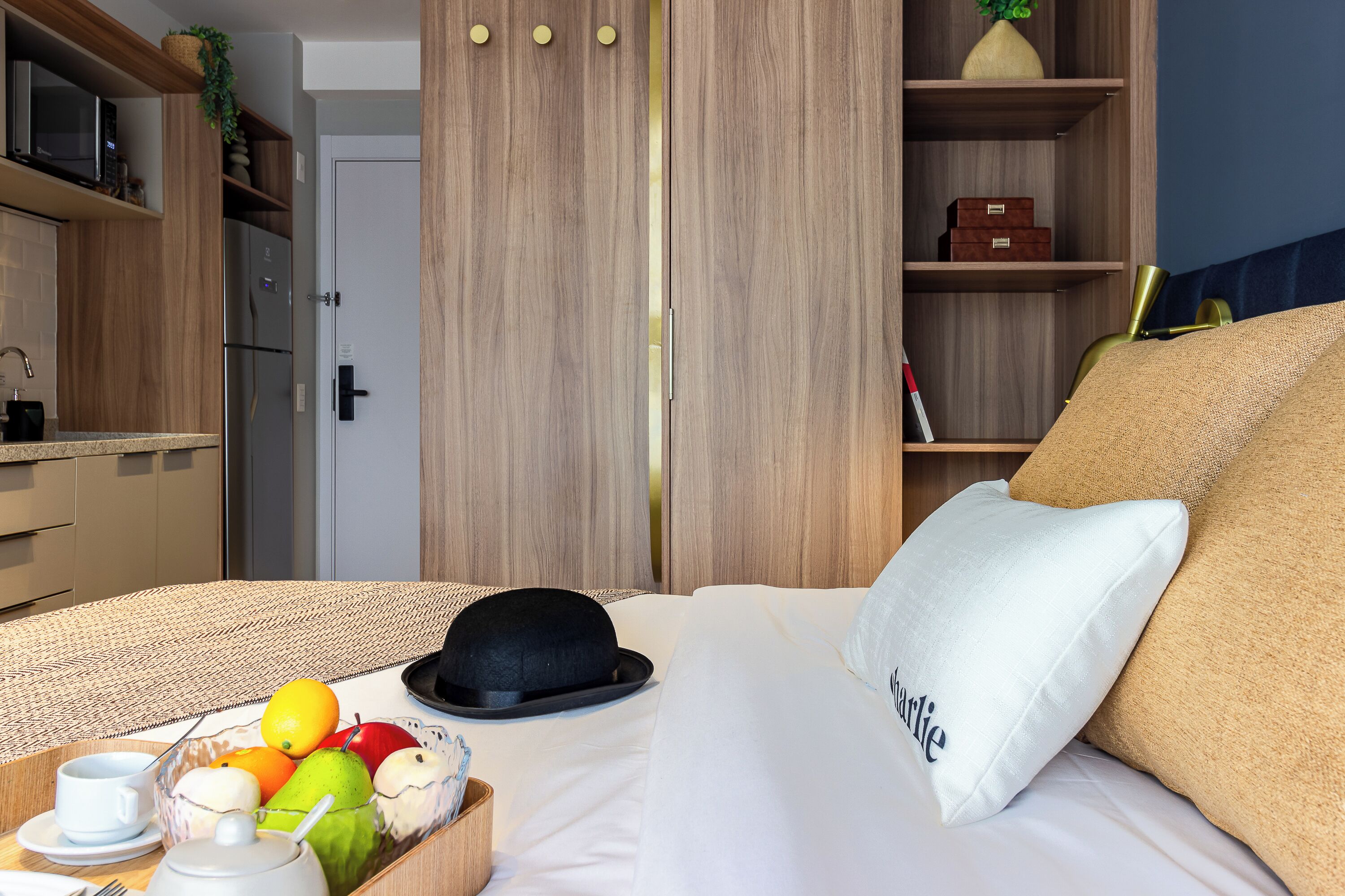 A hotel room with a bed, a bedside table with a fruit bowl, a cup, and a teapot. There is a wardrobe with a mirror and a shelf with decorative items.