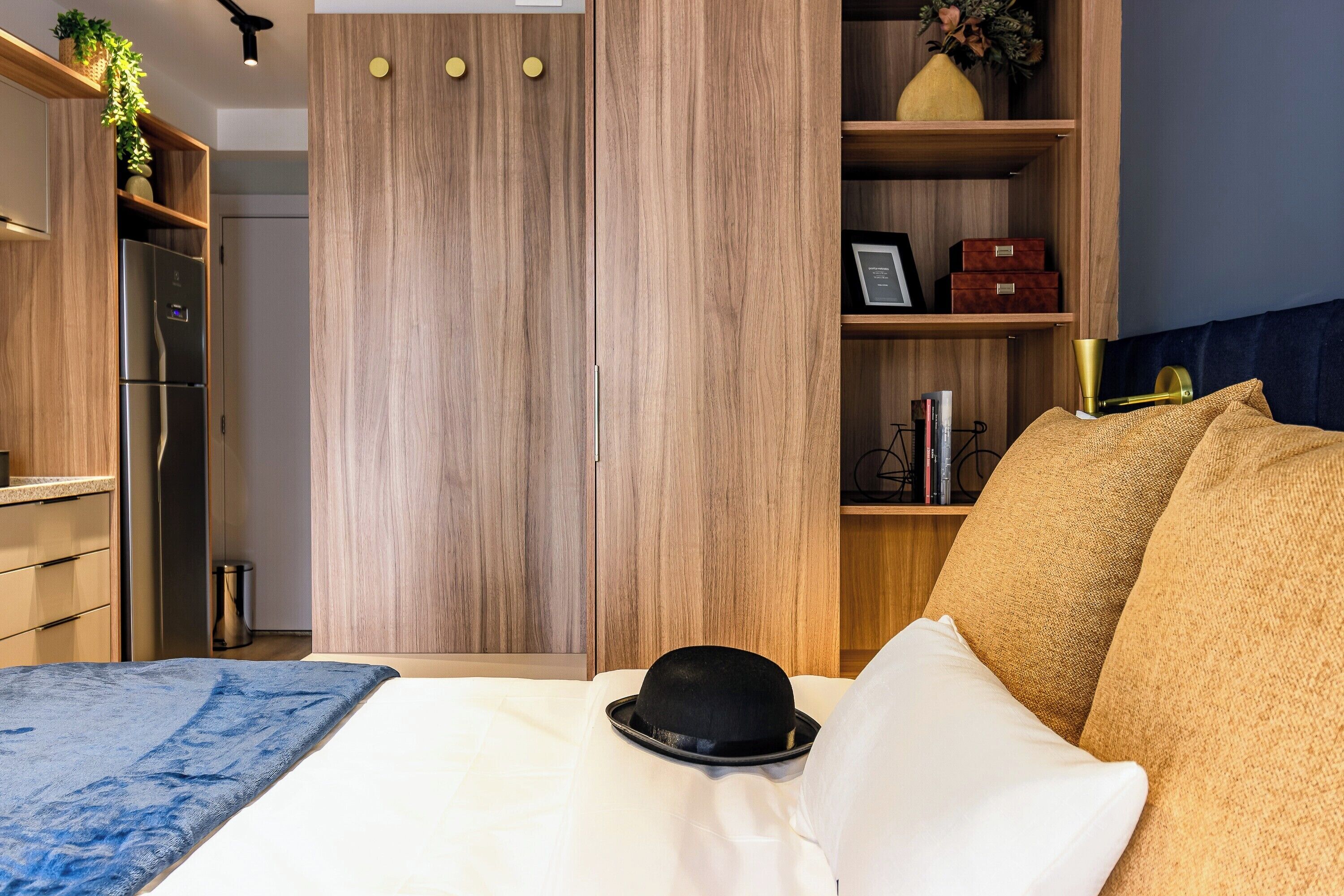 A hotel room with a bed, a wardrobe, a shelf with decorative items, and a small table with a lamp.