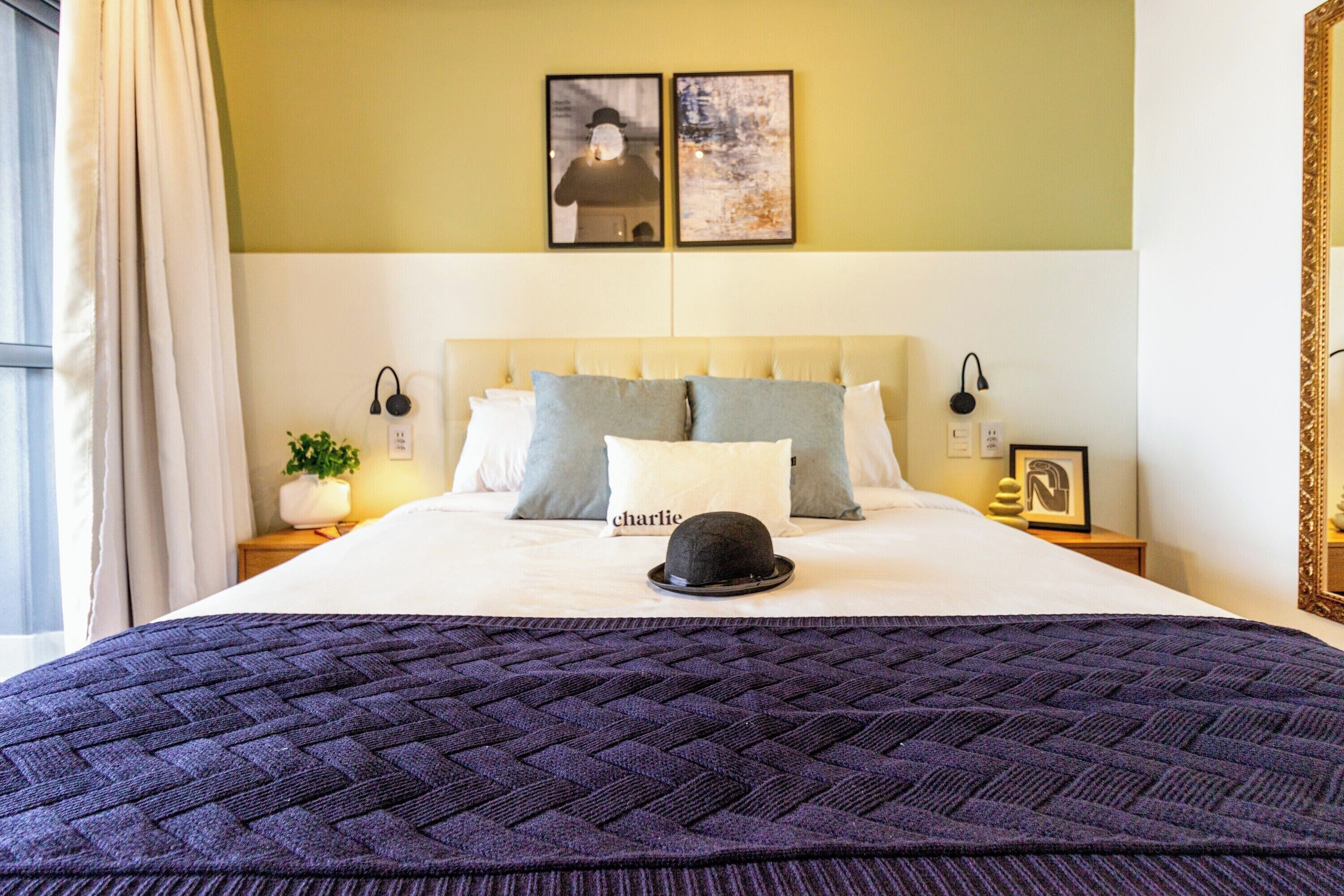 Een netjes opgemaakt bed met een donkere quilt, een hoed op de kussens, en twee ingelijste foto's erboven.