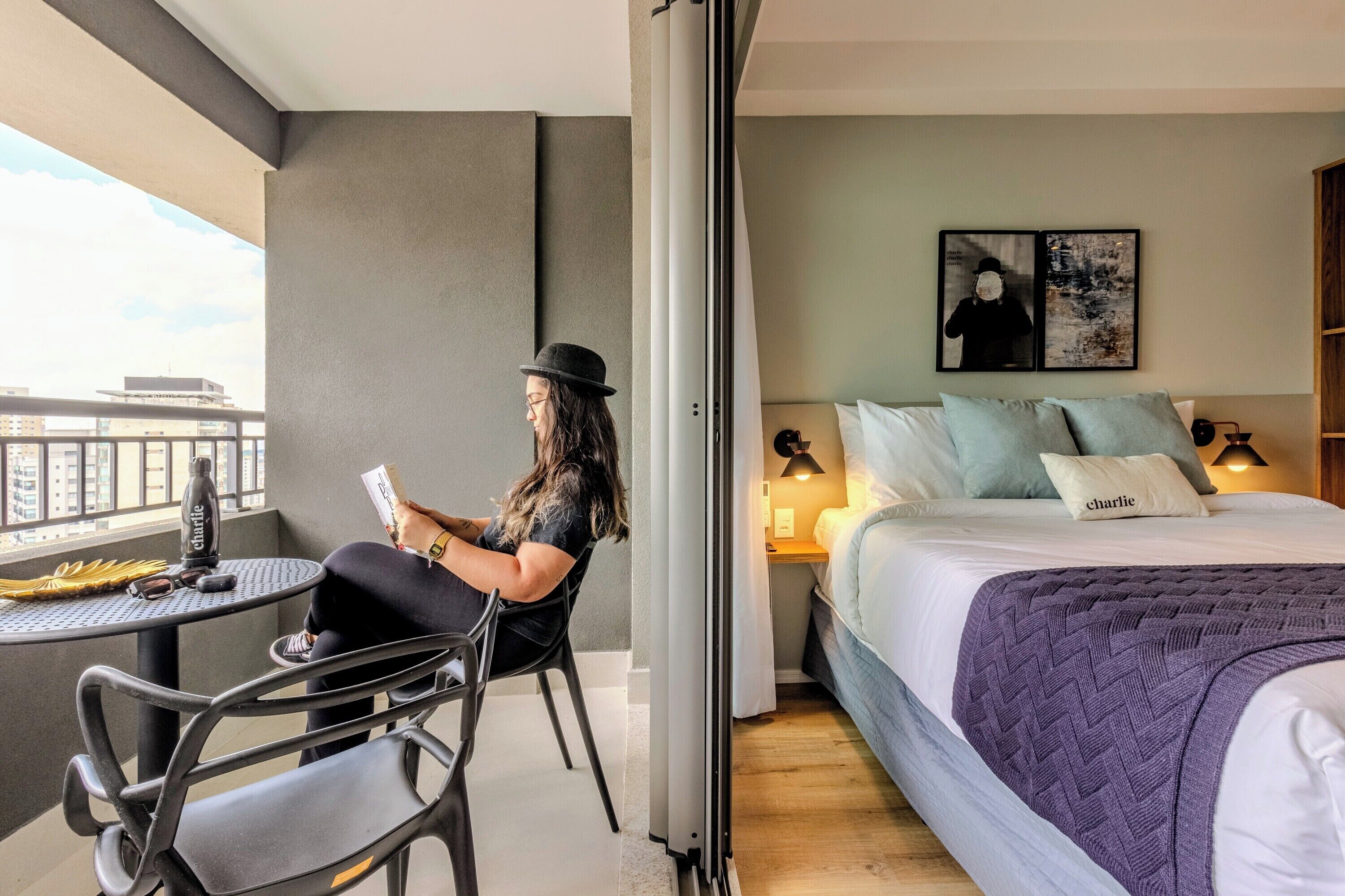 Un balcon avec une table et une chaise, un lit recouvert d’une couverture violette et un mur orné de deux tableaux encadrés.
