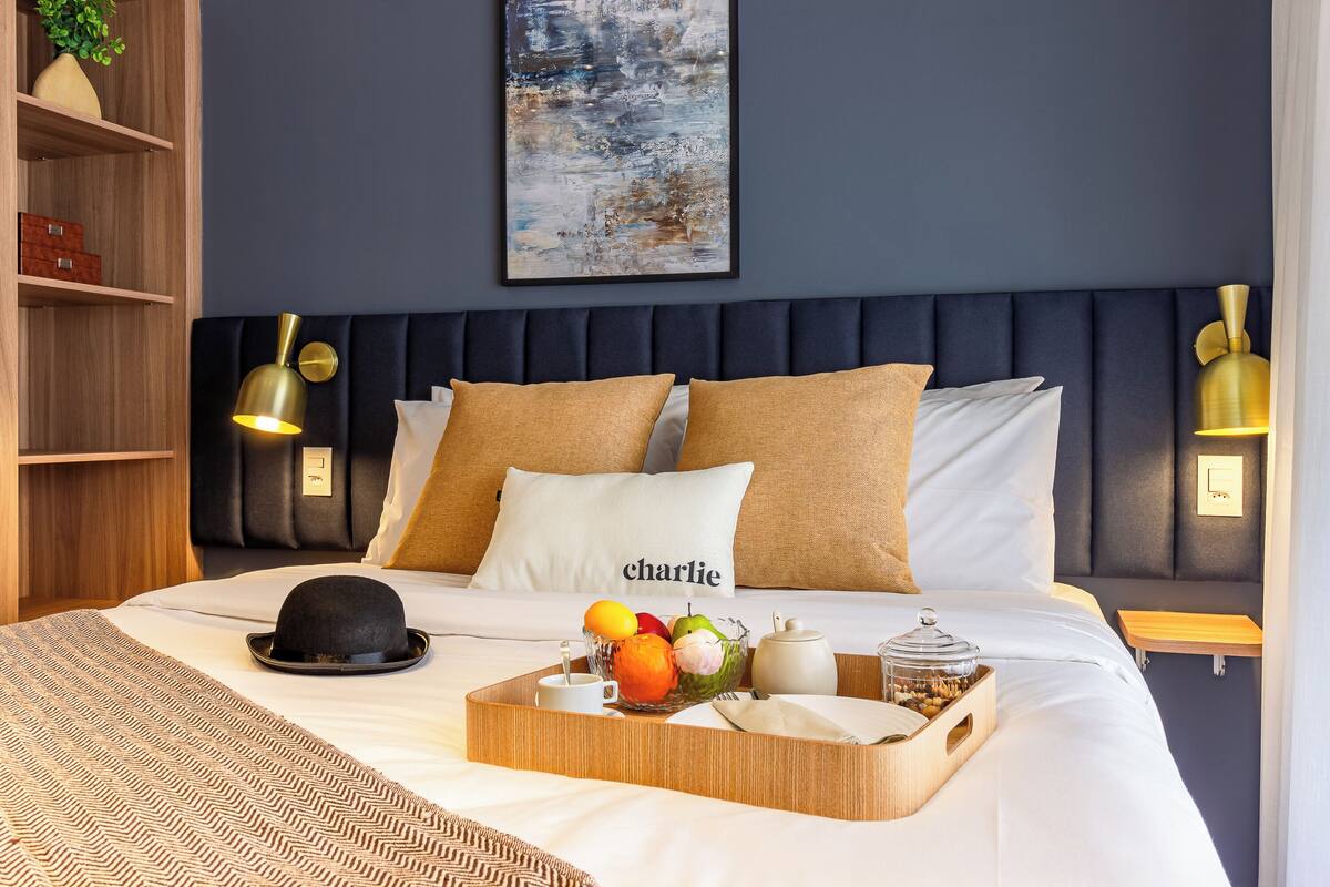 Un letto rifatto con cura, un vassoio di frutta e un cappello, un comodino con una lampada, e una mensola a parete con una pianta.