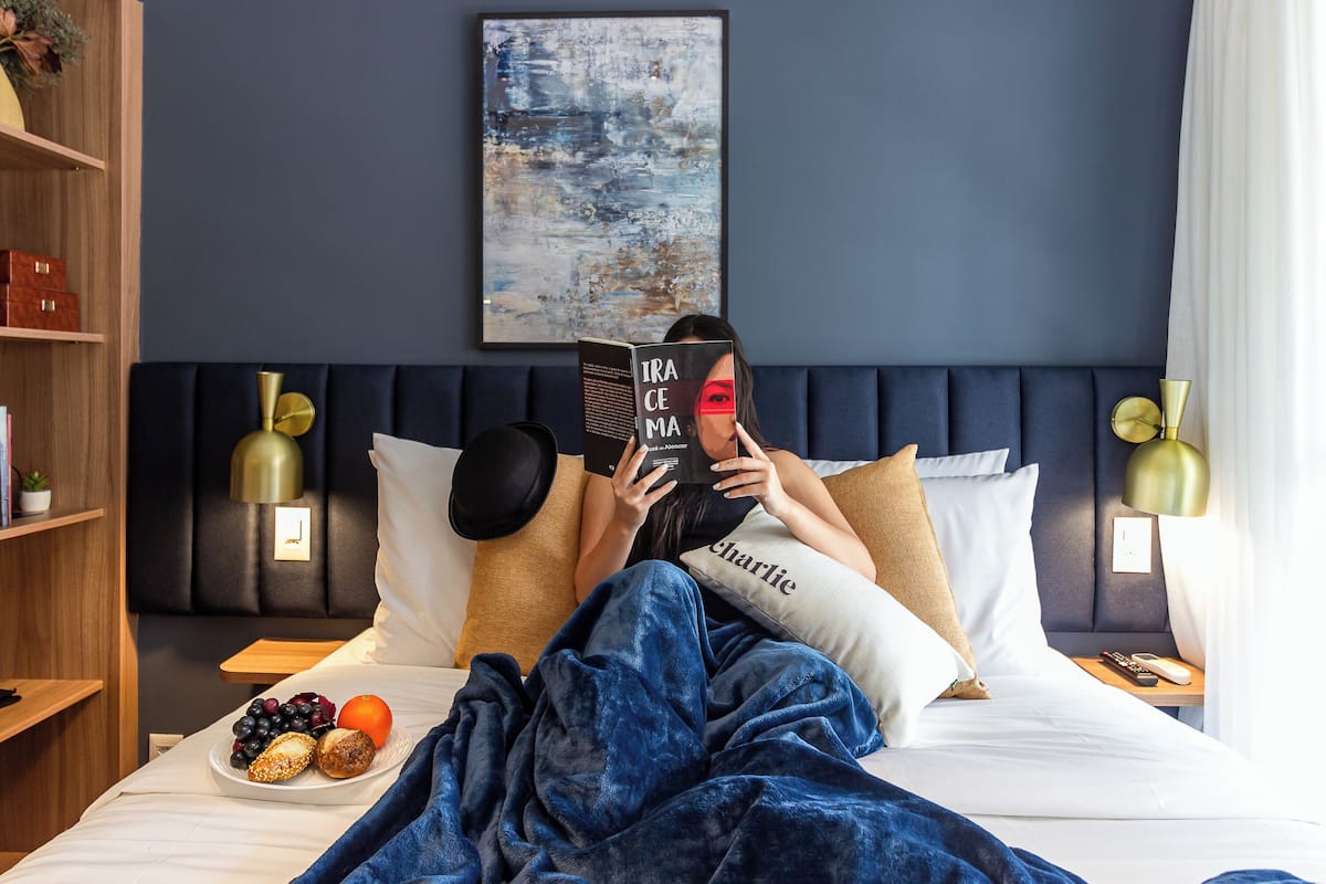 Una camera d'albergo con un letto, un comodino con una lampada, una pianta da notte e un'opera d'arte appesa al muro.