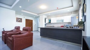 A modern kitchen with white cabinets, a dark countertop, and a microwave. There are three brown massage chairs in the living area. - TCC APARTMENT (Accra)