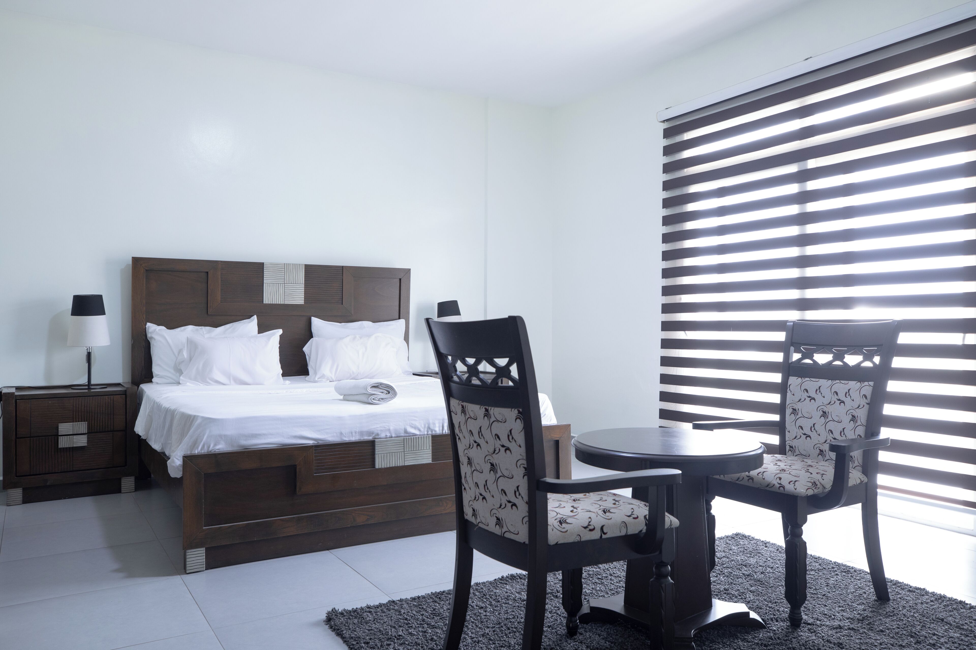 A bedroom with a bed, two chairs, a small table, a nightstand, and a window with blinds.