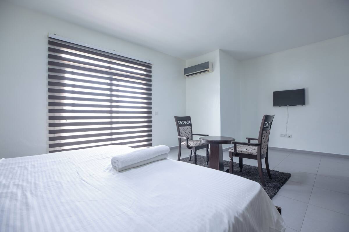 A hotel room with a bed, a small table with two chairs, a TV mounted on the wall, and a window with blinds.