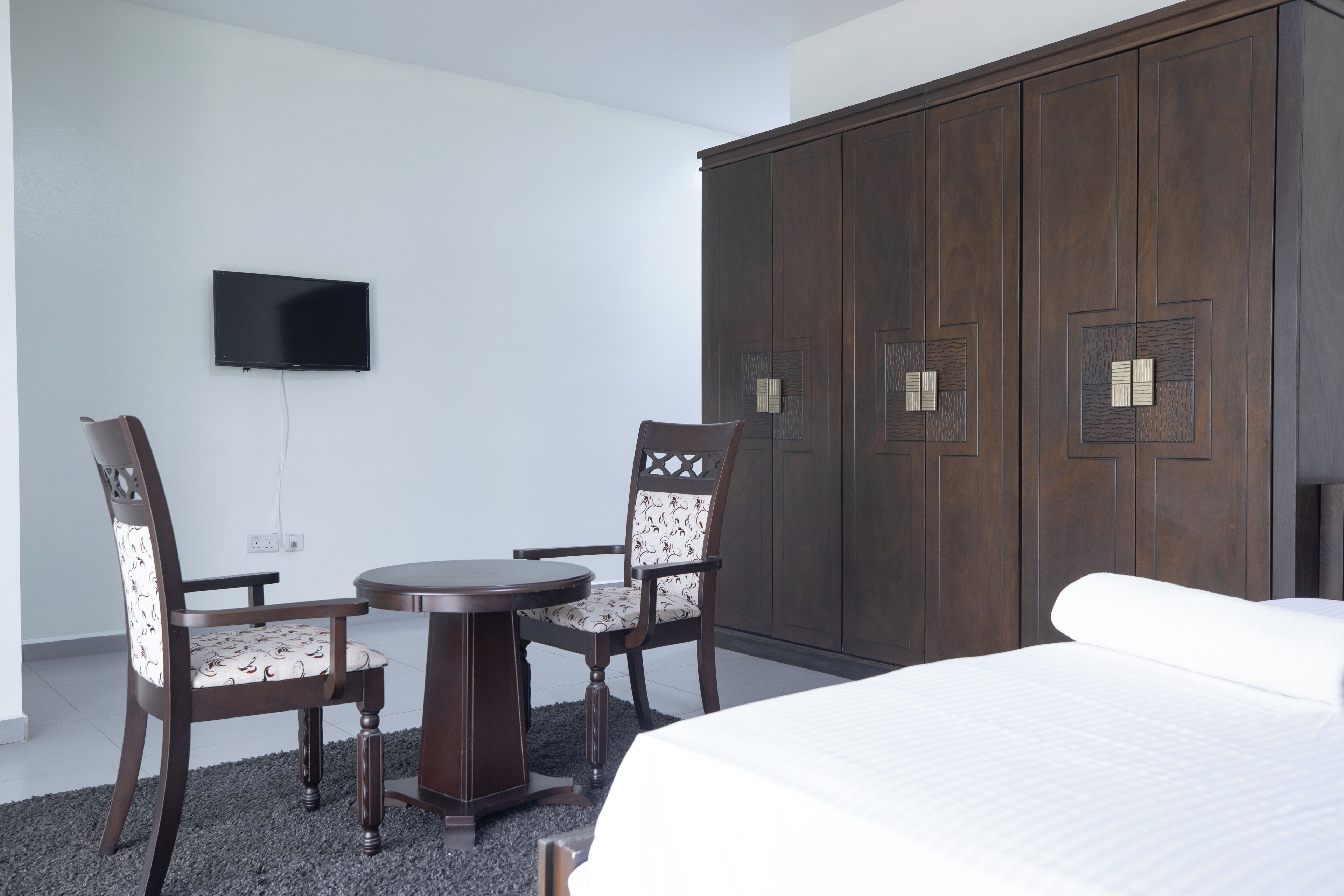 A bedroom with a bed, a TV mounted on the wall, a wardrobe, and two chairs around a small table.
