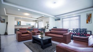A modern living room with leather sofas, a coffee table, and a kitchen in the background. - TCC APARTMENT (Accra)