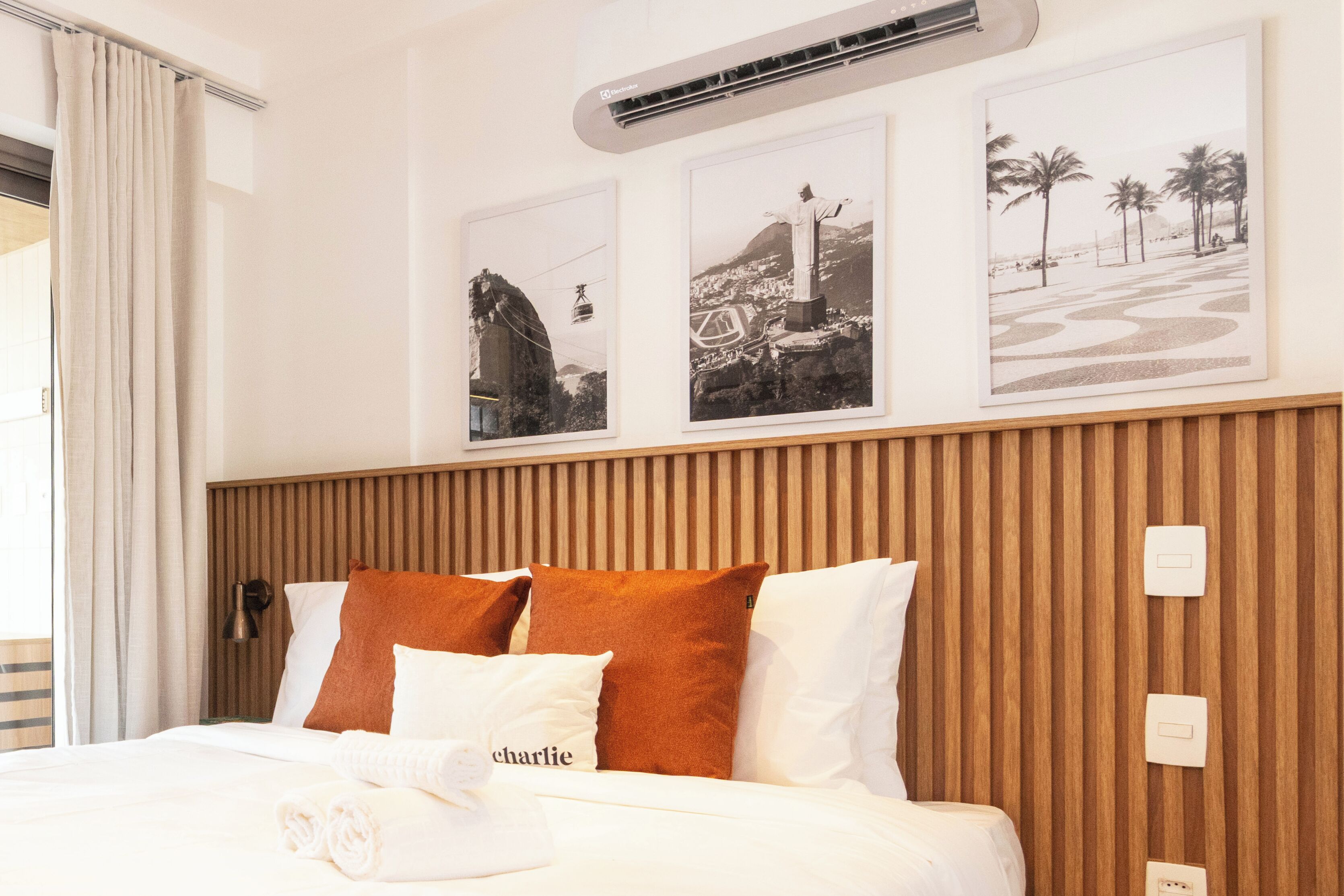 A hotel room with a bed, a wall-mounted air conditioner, and three framed pictures depicting famous landmarks.