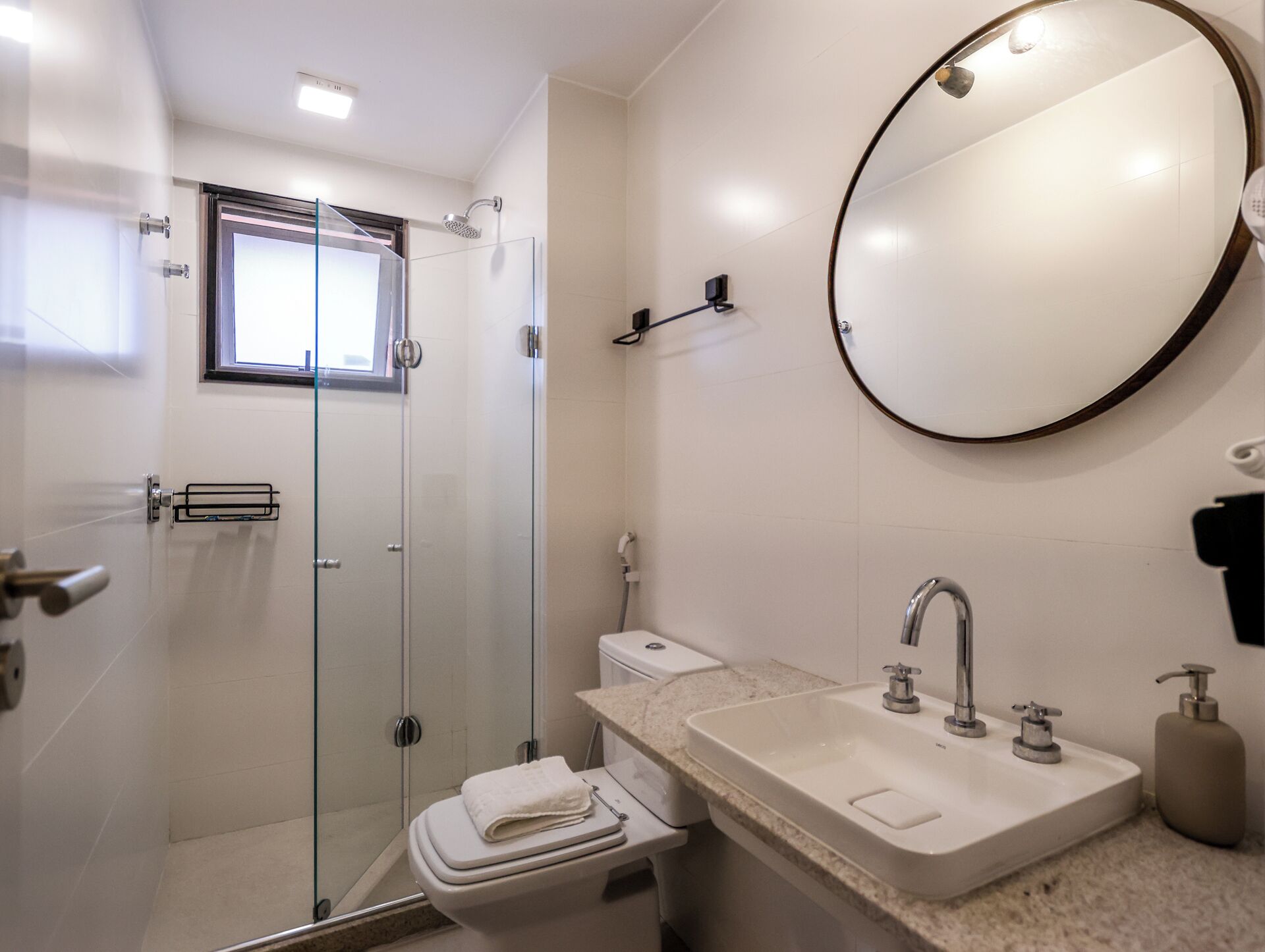 A modern bathroom with a glass shower enclosure, a toilet, a sink with a marble countertop, and a round mirror.