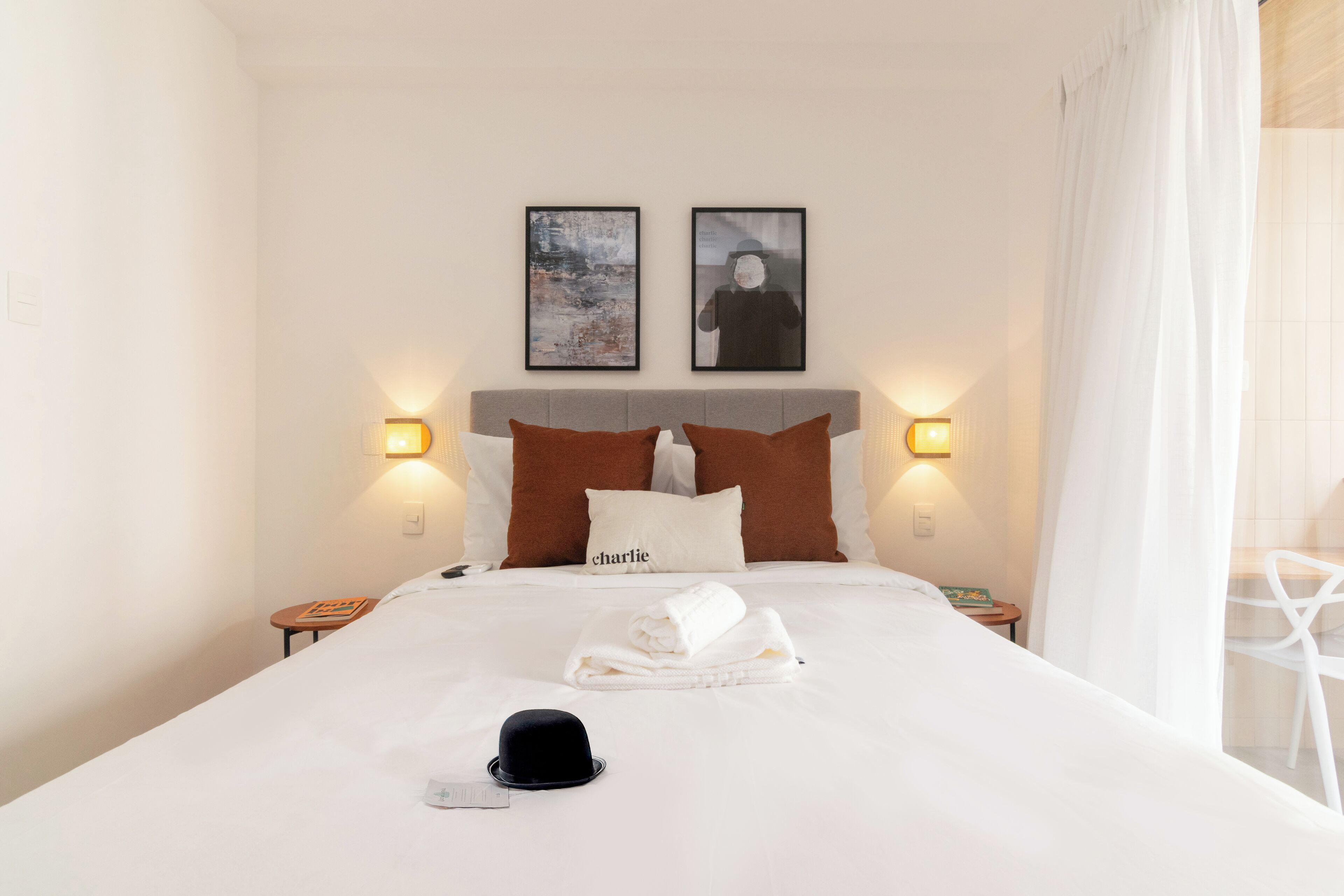 A neatly made bed with a white comforter, a folded towel, and a black hat on the bed. There are two pillows, one with the name "Charlie" and another with a blurred face. Two framed pictures are hung above the bed, and there are two wall-mounted lamps on either side of the bed.