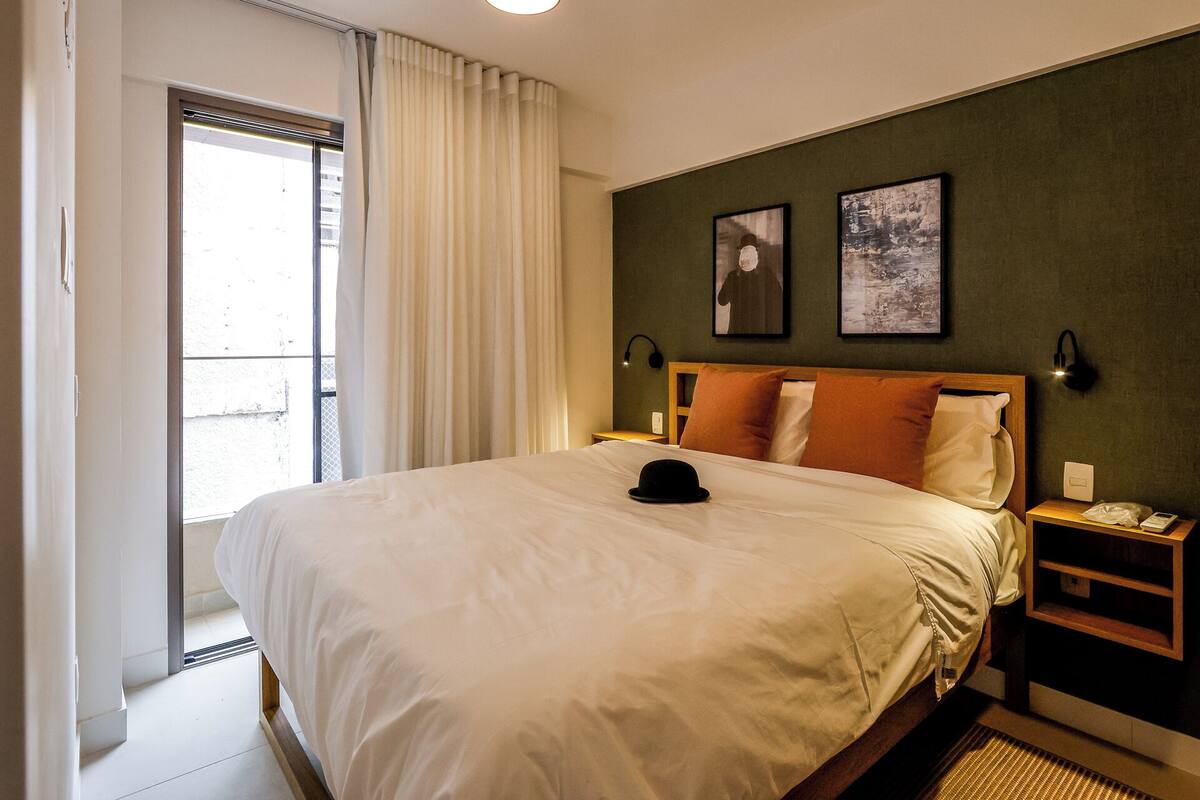 A hotel room with a large bed, a nightstand, two wall-mounted lamps, and a window with curtains.