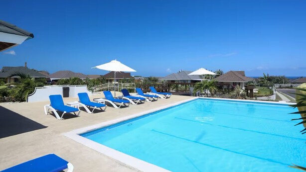 A swimming pool with blue lounge chairs and umbrellas, surrounded by houses with sloped roofs. - Tropical Tranquility in Jamaica (Richmond Estate)