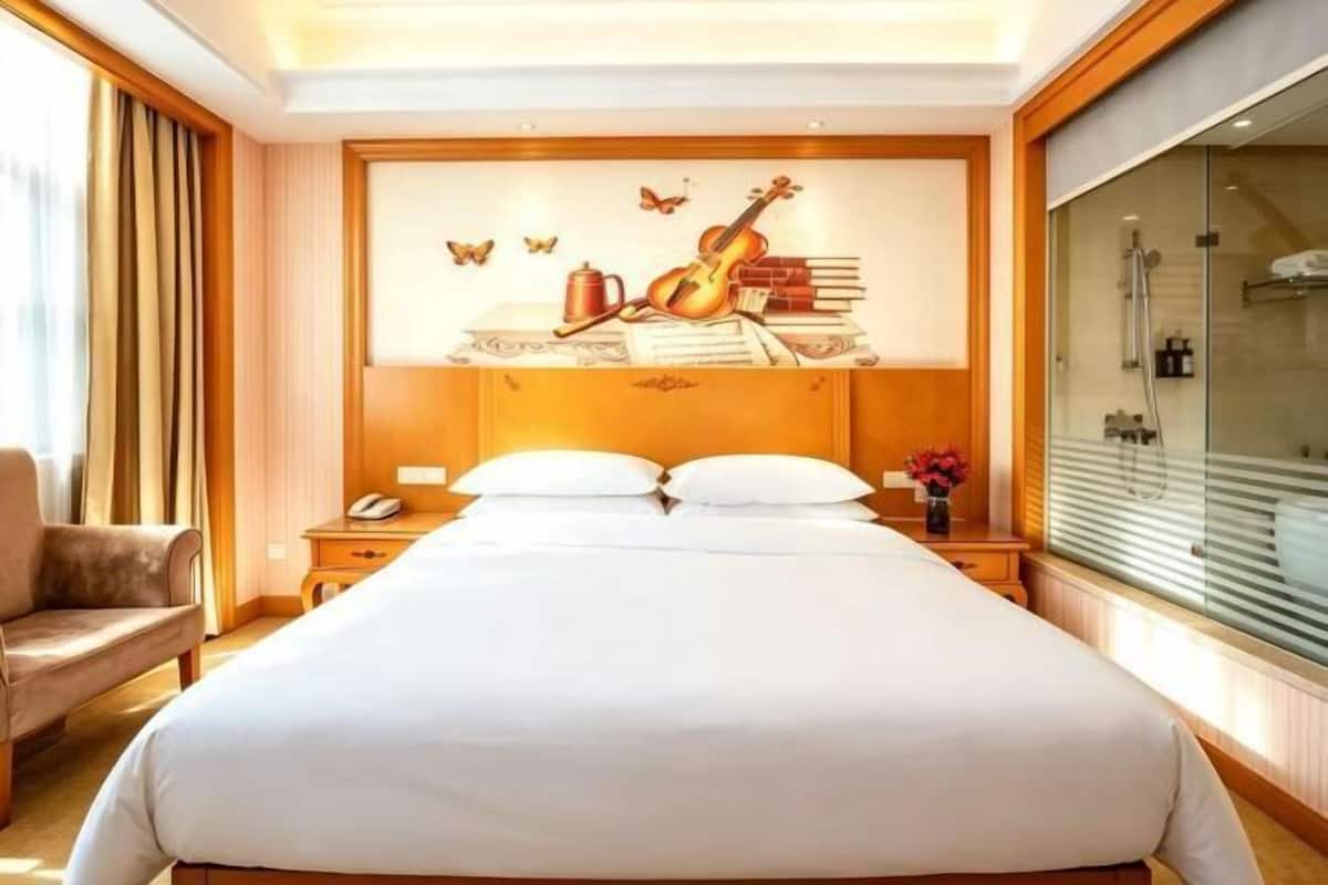 A hotel room with a large bed, a nightstand, a chair, a wall with a guitar and butterflies mural, and a window with a view.