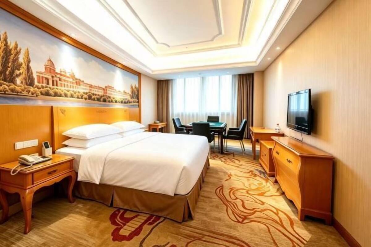 A hotel room with a large bed, a desk, a television, and a painting on the wall.