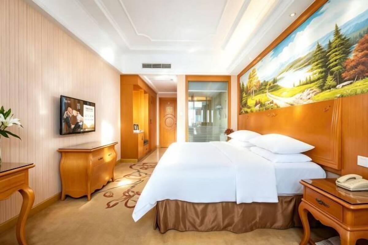 A hotel room with a large bed, a desk, a chair, a painting, and a view of a forest.