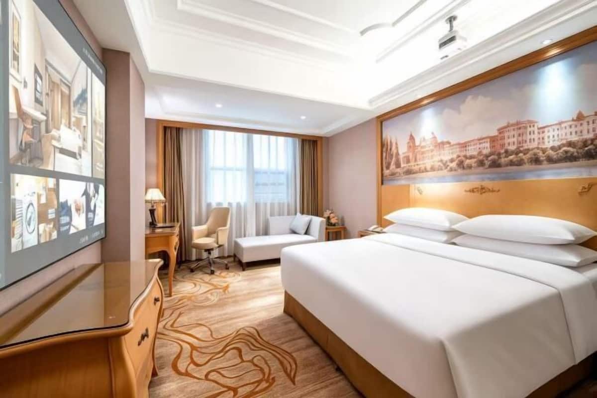 A hotel room with a large bed, a desk, a chair, a TV, and a painting on the wall.