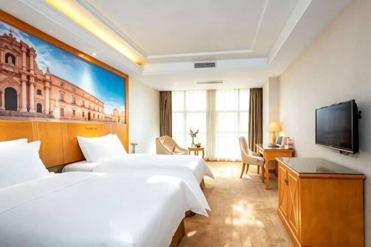 A hotel room with two beds, a TV, a desk, a chair, and a painting of a building on the wall.