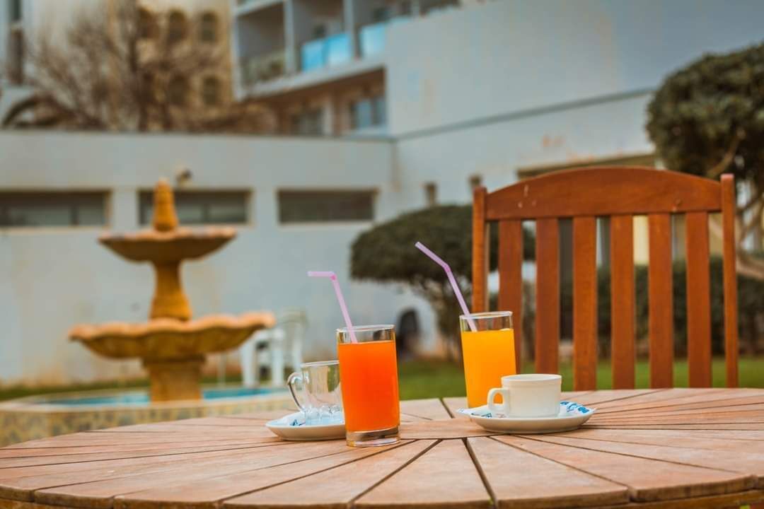 A wooden table with two glasses of orange juice and a white cup, set against a building with a fountain in the background.