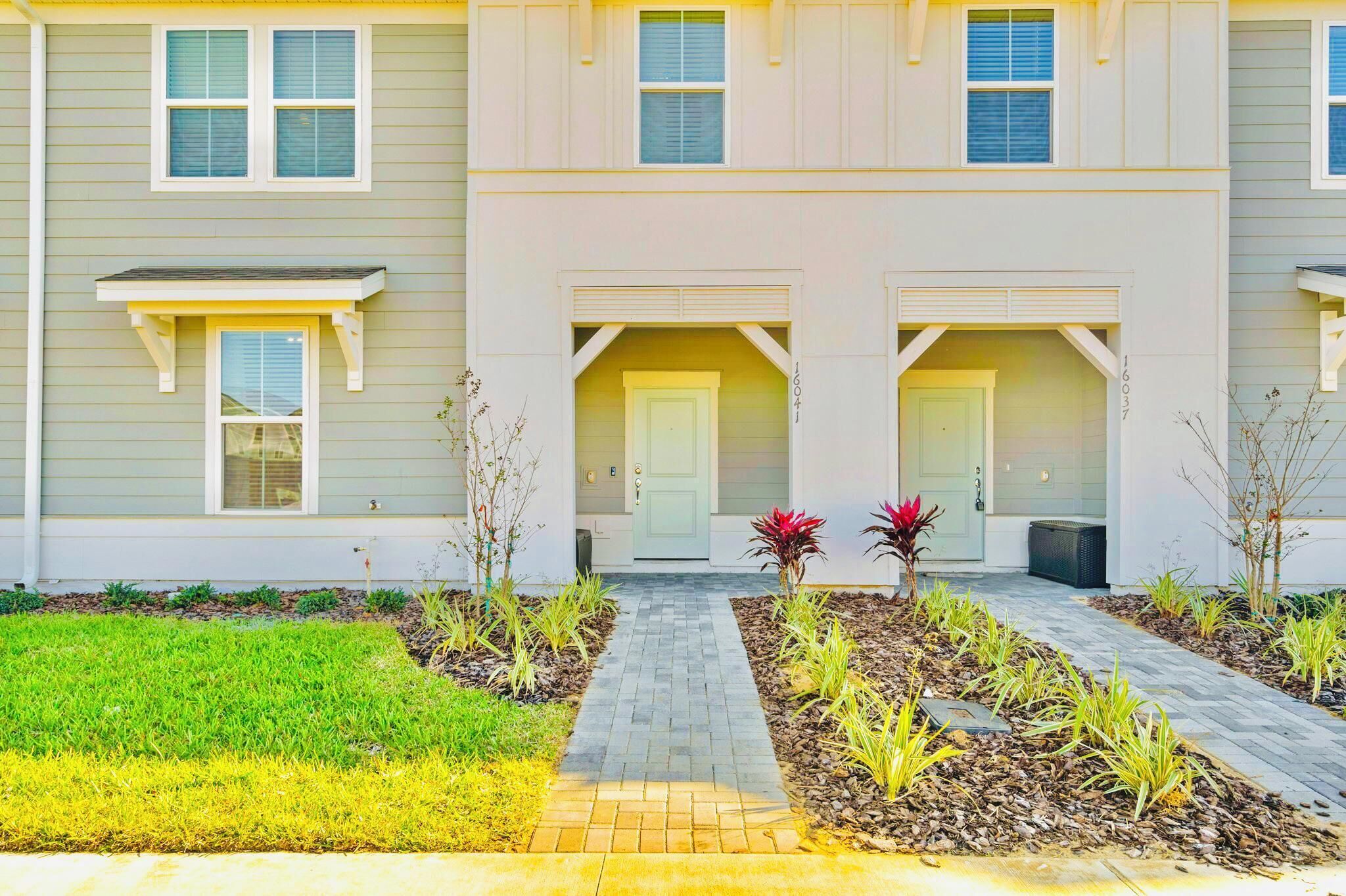 A two-story residential building with a front porch, a central door, and a small garden area with plants and a pathway.
