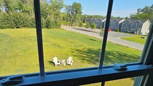A view through a window of a residential area with a lawn, two Adirondack chairs, and a wooden deck. - Acadia Villas!  5B Hunter's Way:  A private neighborhood nestled in the woods. (Trenton)