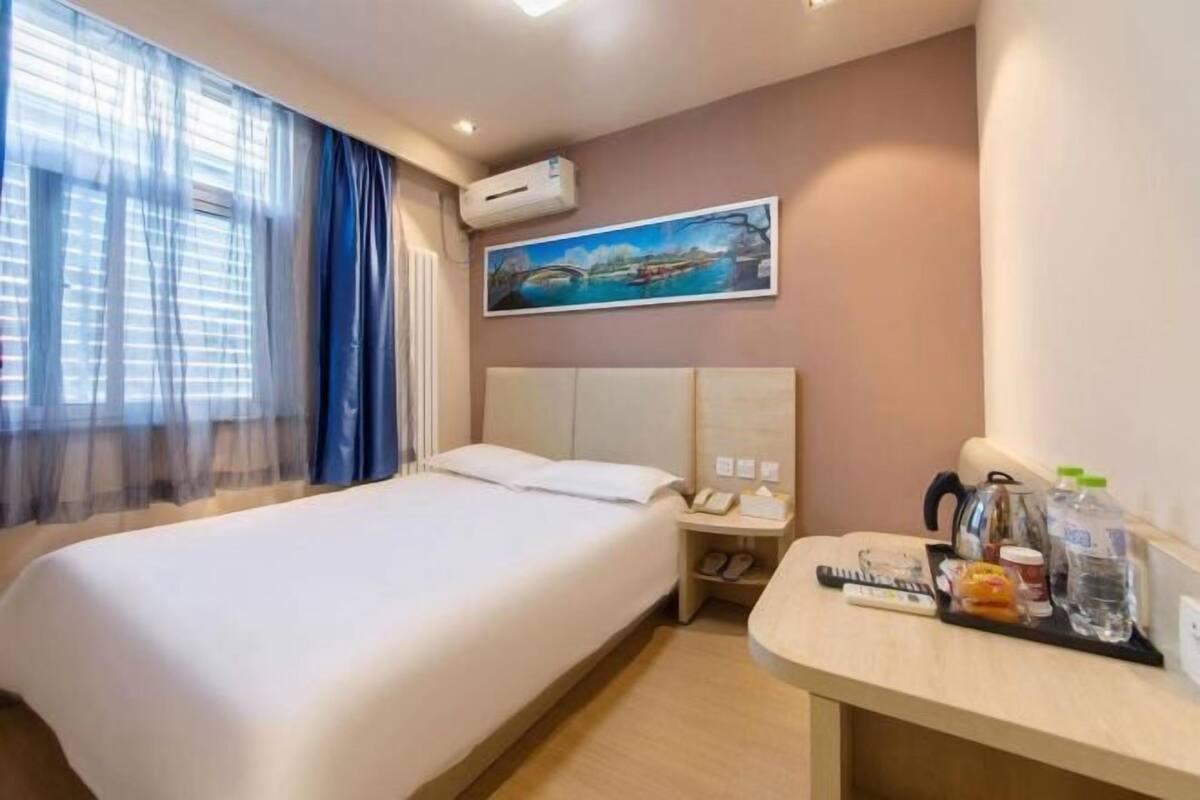 A hotel room with a bed, a nightstand, a painting, and a desk with a kettle and water bottles.