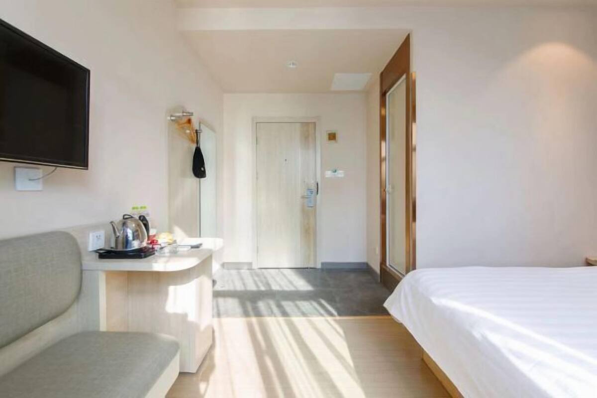 A hotel room with a bed, a sofa, a TV, a desk with a kettle, and a door leading to another room.